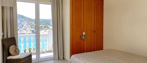 Um quarto com uma cama, uma cadeira de vime, uma porta de madeira e uma varanda com vista para as colinas.