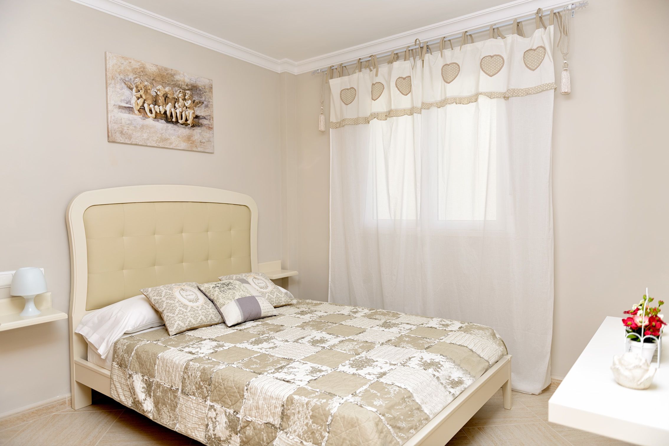 Un dormitorio con cama, mesita de noche, cortinas translúcidas en la ventana y una obra de arte colgada en la pared.