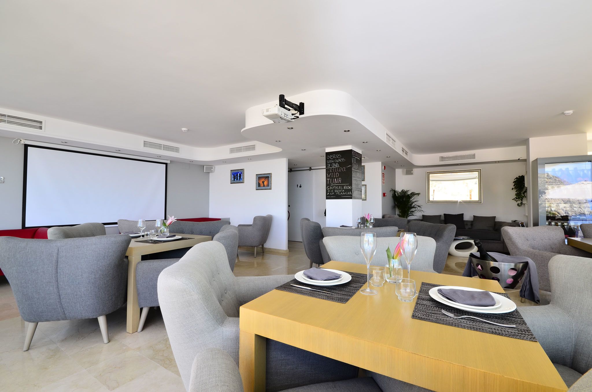 A modern living room with a dining area, grey sofas, a wooden dining table, and a large screen.