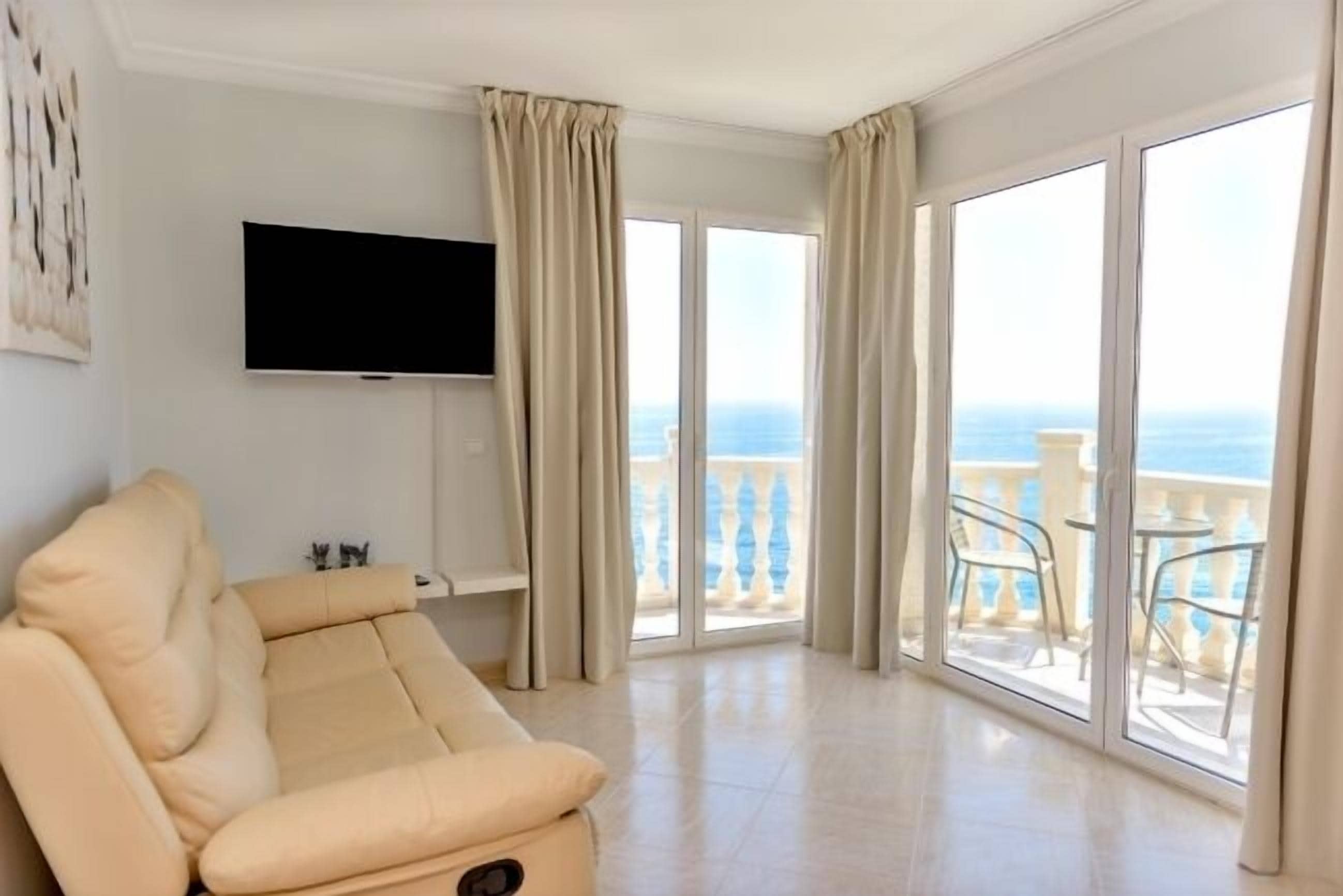 A spacious living room with a beige sofa, a flat-screen TV mounted on the wall, and large sliding glass doors leading to a balcony with a view of the sea.