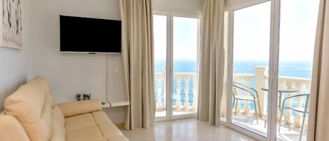 A spacious living room with a beige sofa, a flat-screen TV mounted on the wall, and large sliding glass doors leading to a balcony with a view of the sea.