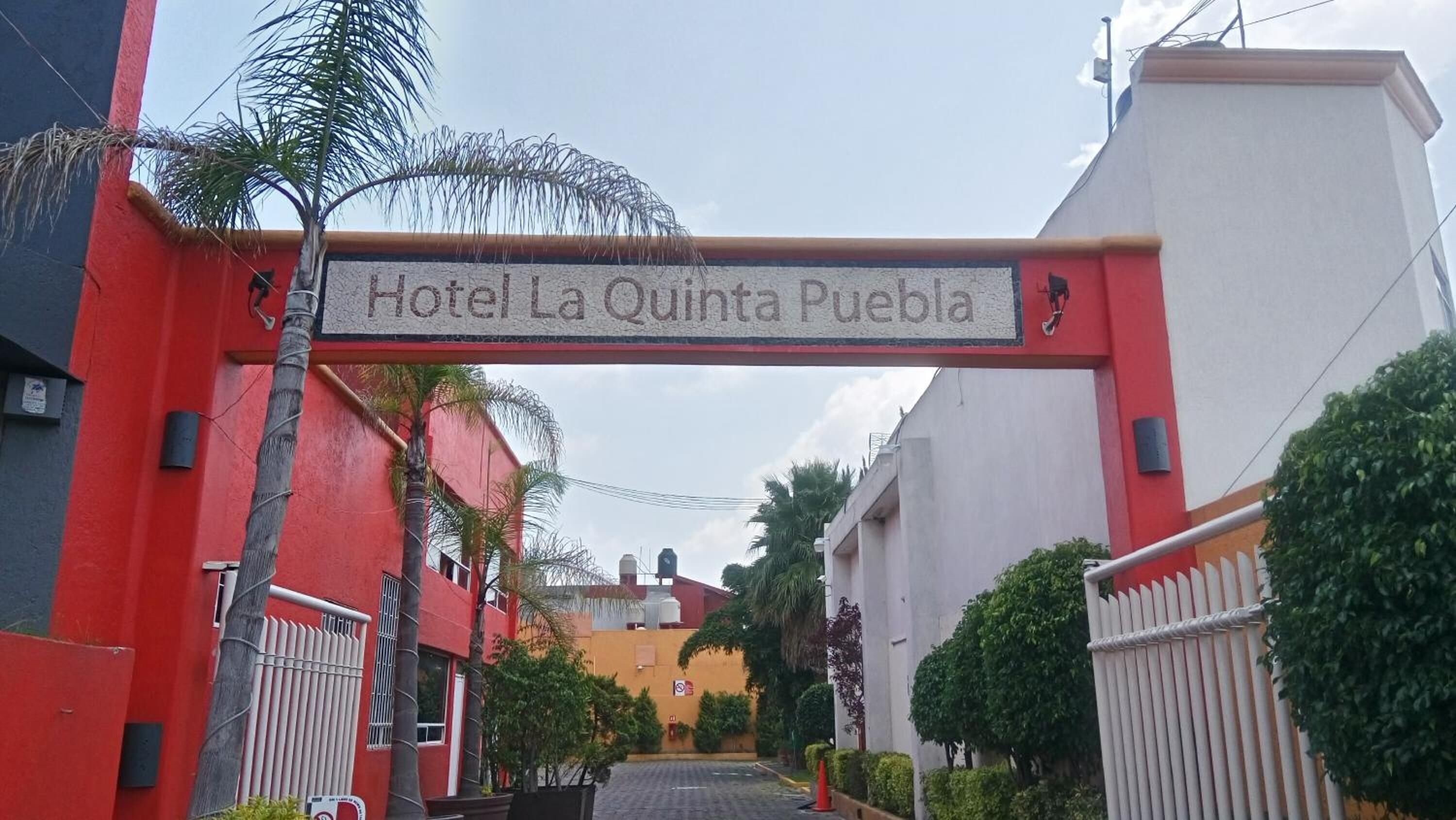 Ingången till Hotel La Quinta Puebla, med en tydlig skylt, en uppfart med grind och omgivande grönska.