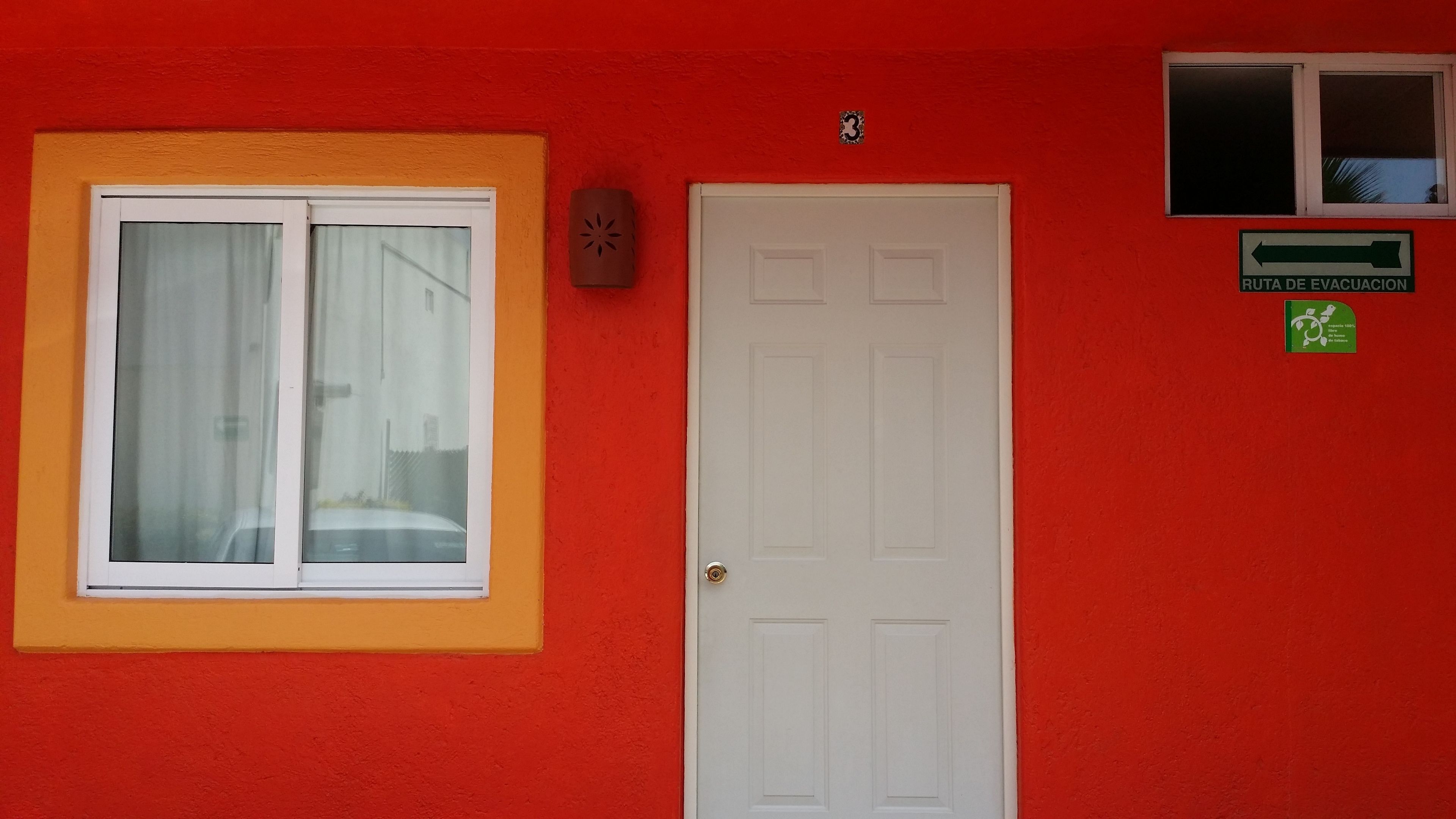 Un edificio rojo con puerta blanca, una ventana con cortina y un letrero que indica una ruta de evacuación.
