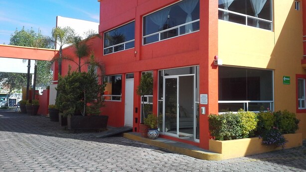Un edificio de dos pisos con fachada roja y amarilla, ventanas amplias y un área de entrada pavimentada con plantas en macetas.