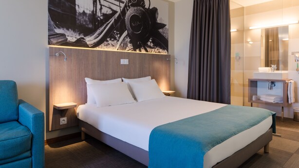 A modern hotel room with a large bed, a side table, a chair, a bathroom with a mirror, and a wall-mounted artwork.