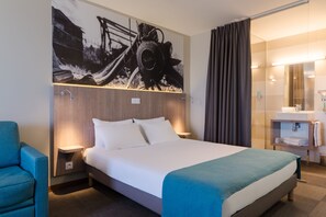 Habitación de hotel moderna con una cama grande, una mesita de noche, una silla, un baño con espejo y una obra de arte colgada en la pared.