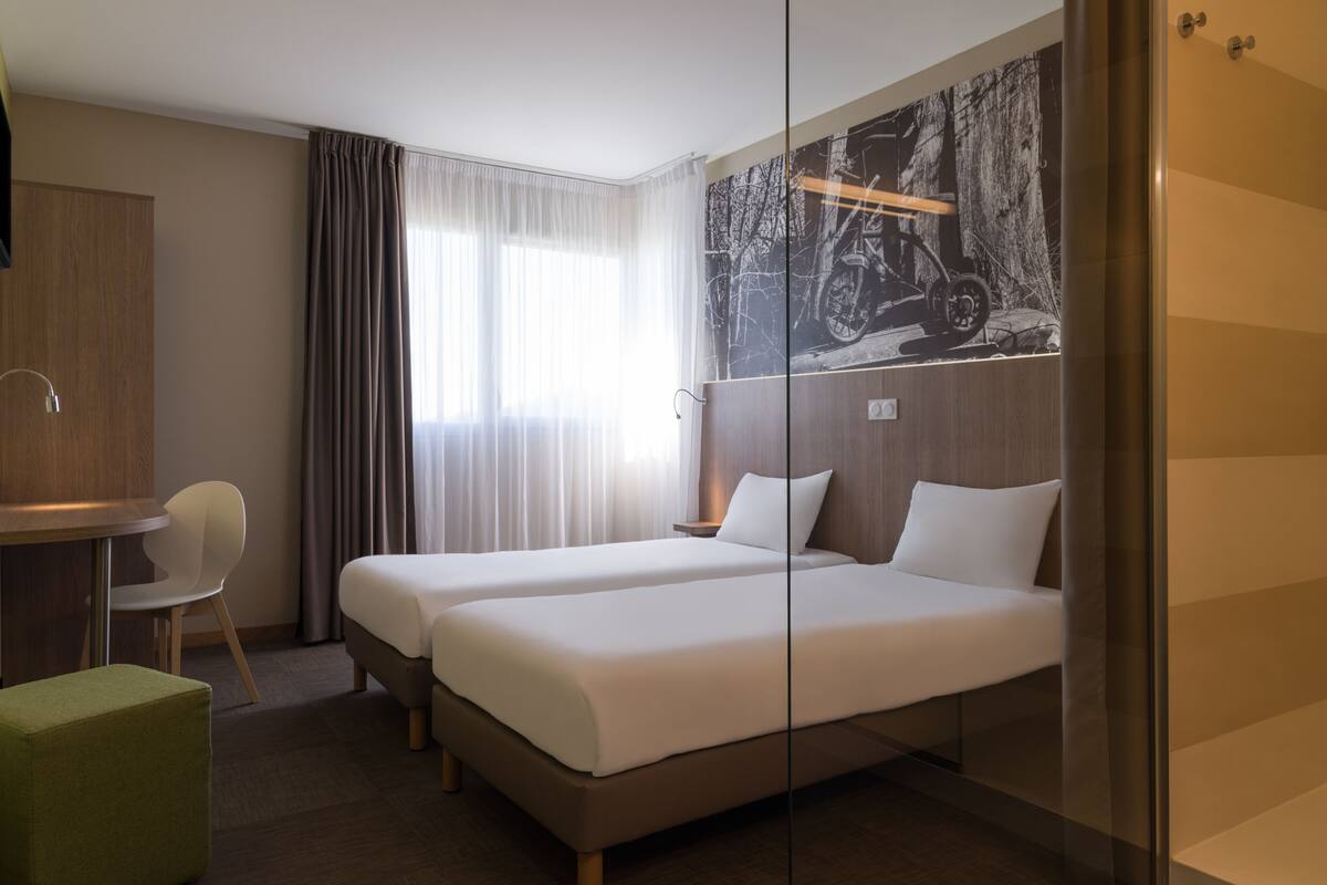 Habitación de hotel con dos camas individuales, un escritorio con silla, un otoman verde y una obra de arte grande colgada en la pared.