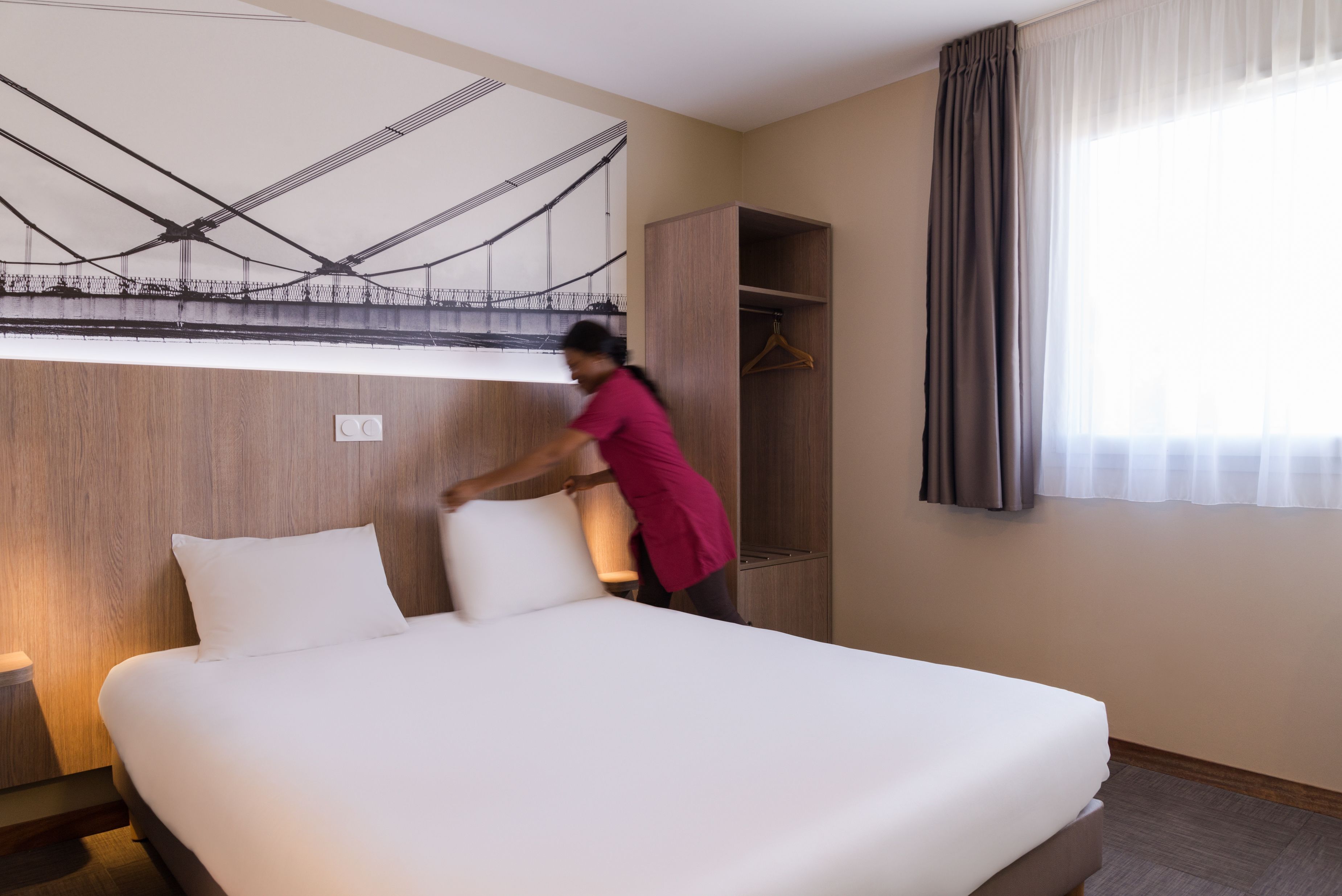 A hotel room with a neatly made bed, a wooden wardrobe, a coat hanger, and a large window with curtains.