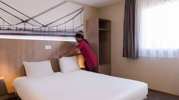 A hotel room with a neatly made bed, a wooden wardrobe, a coat hanger, and a large window with curtains.