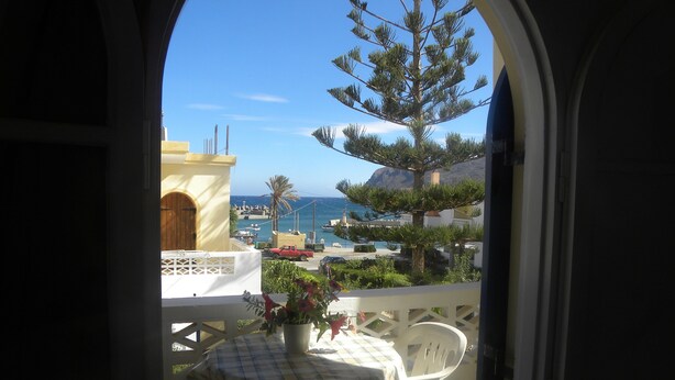 A balcony with a table and chairs, overlooking a coastal area with buildings, a palm tree, and a view of the sea. - Villa Thalia (Agios Nikolaos)