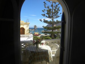 A balcony with a table and chairs, overlooking a coastal area with buildings, a palm tree, and a view of the sea. - Villa Thalia (Agios Nikolaos)