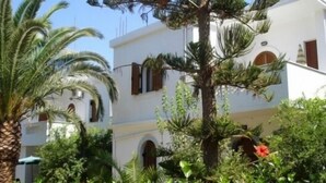 A white building with multiple balconies, surrounded by lush greenery and palm trees. - Villa Thalia (Agios Nikolaos)