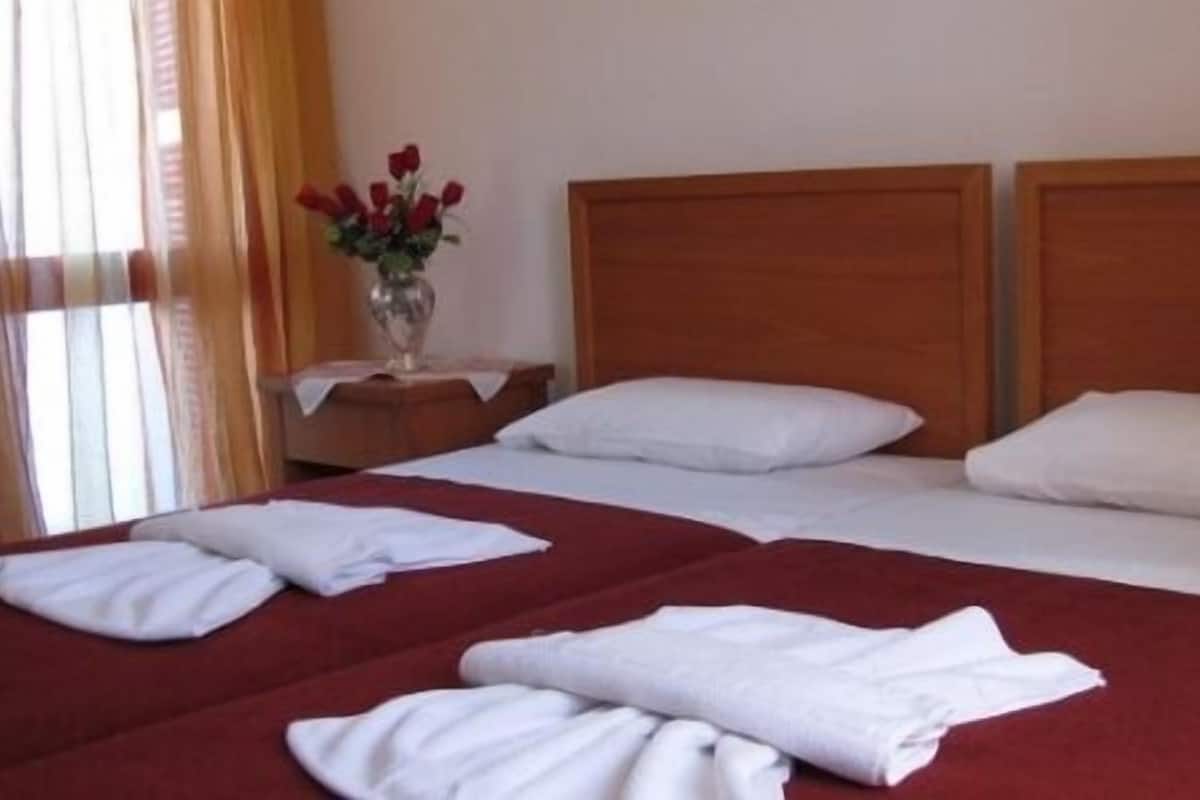 Habitación de hotel con dos camas, cada una con ropa de cama blanca y colchas rojas. Se observa un cabecero de madera, una mesita de noche con un jarrón de flores rojas y una ventana con cortinas.