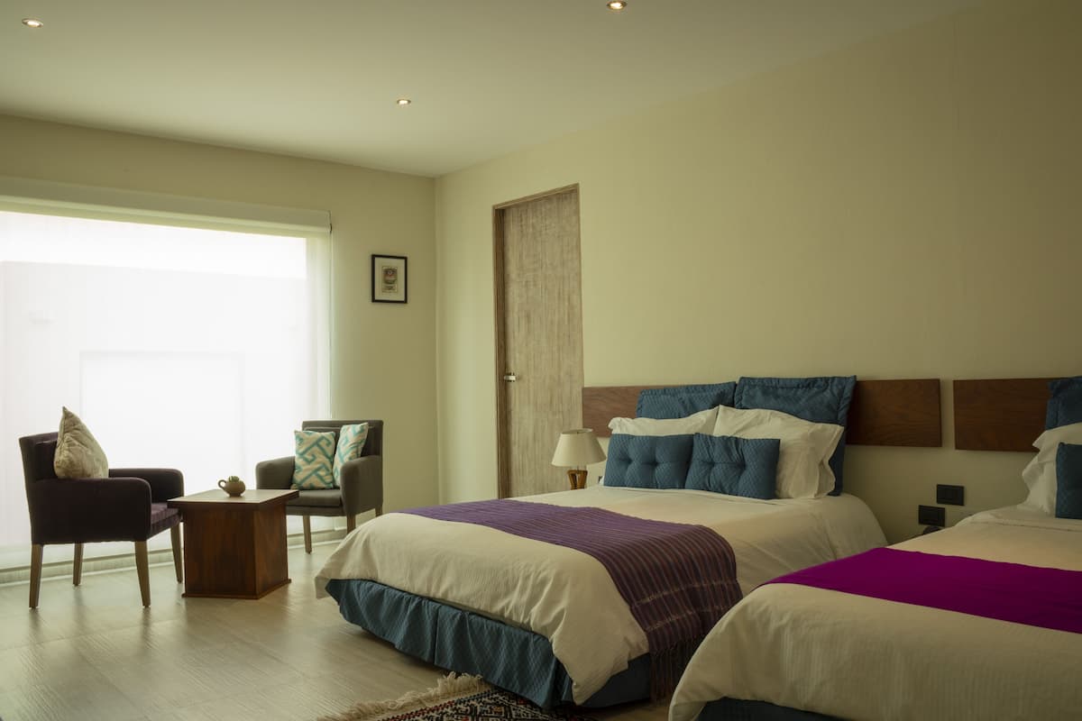 Une chambre d’hôtel avec deux lits, un coin salon comprenant une chaise et une petite table, ainsi qu’une grande fenêtre laissant entrer la lumière naturelle.