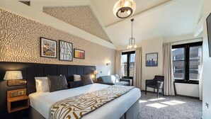 A hotel room with a large bed, a headboard, a nightstand, a desk, and a chair. There are framed pictures on the wall, a window with curtains, and a ceiling light. - The Abel Heywood (Manchester)