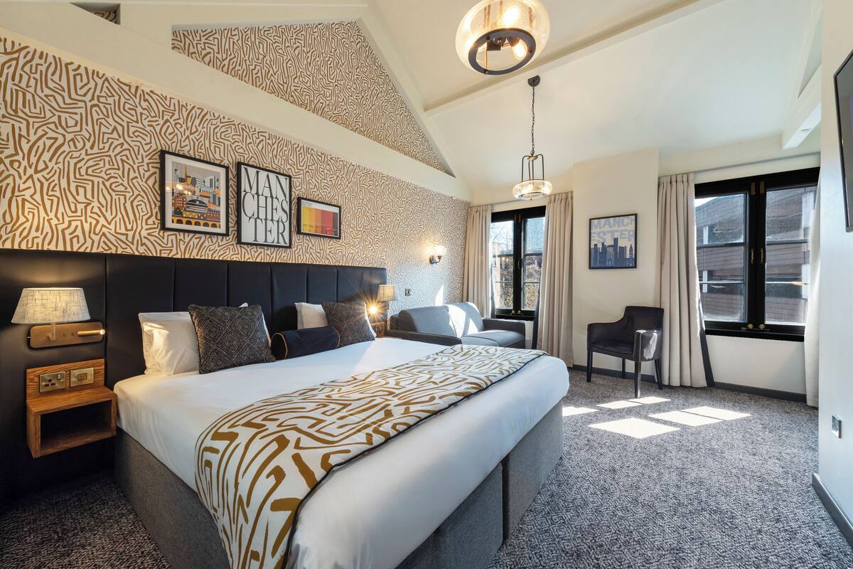 A hotel room with a large bed, a headboard, a nightstand, a desk, and a chair. There are framed pictures on the wall, a window with curtains, and a ceiling light.