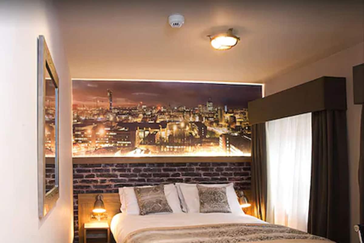 A hotel room with a cityscape view, a bed with a dark headboard, a nightstand with a lamp, and a window with curtains.