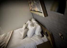 Una cama bien hecha con sábanas y almohadones blancos, una mesita de noche con lámpara, y una obra de arte colgada en la pared sobre la cama.