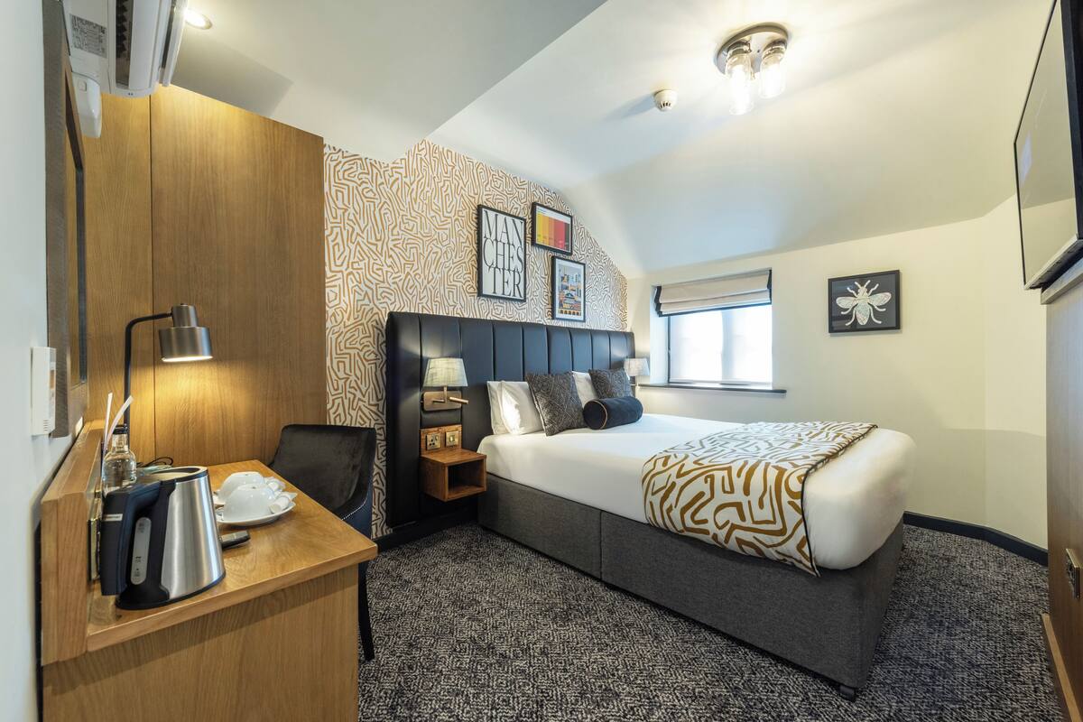 A hotel room with a bed, a desk with a kettle, a chair, a nightstand, and a wall with patterned wallpaper and framed pictures.