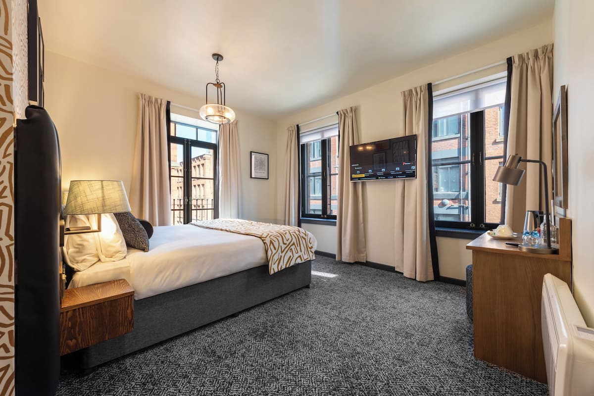 A hotel room with a large bed, a desk with a lamp, a TV mounted on the wall, and a window with curtains.