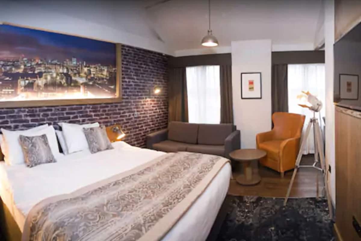 A hotel room with a large bed, a sofa, a chair, a small table, a lamp, and a cityscape painting on the wall.