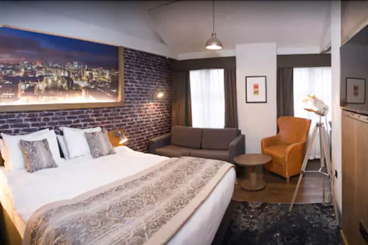 A hotel room with a large bed, a sofa, a chair, a small table, a lamp, and a cityscape painting on the wall.