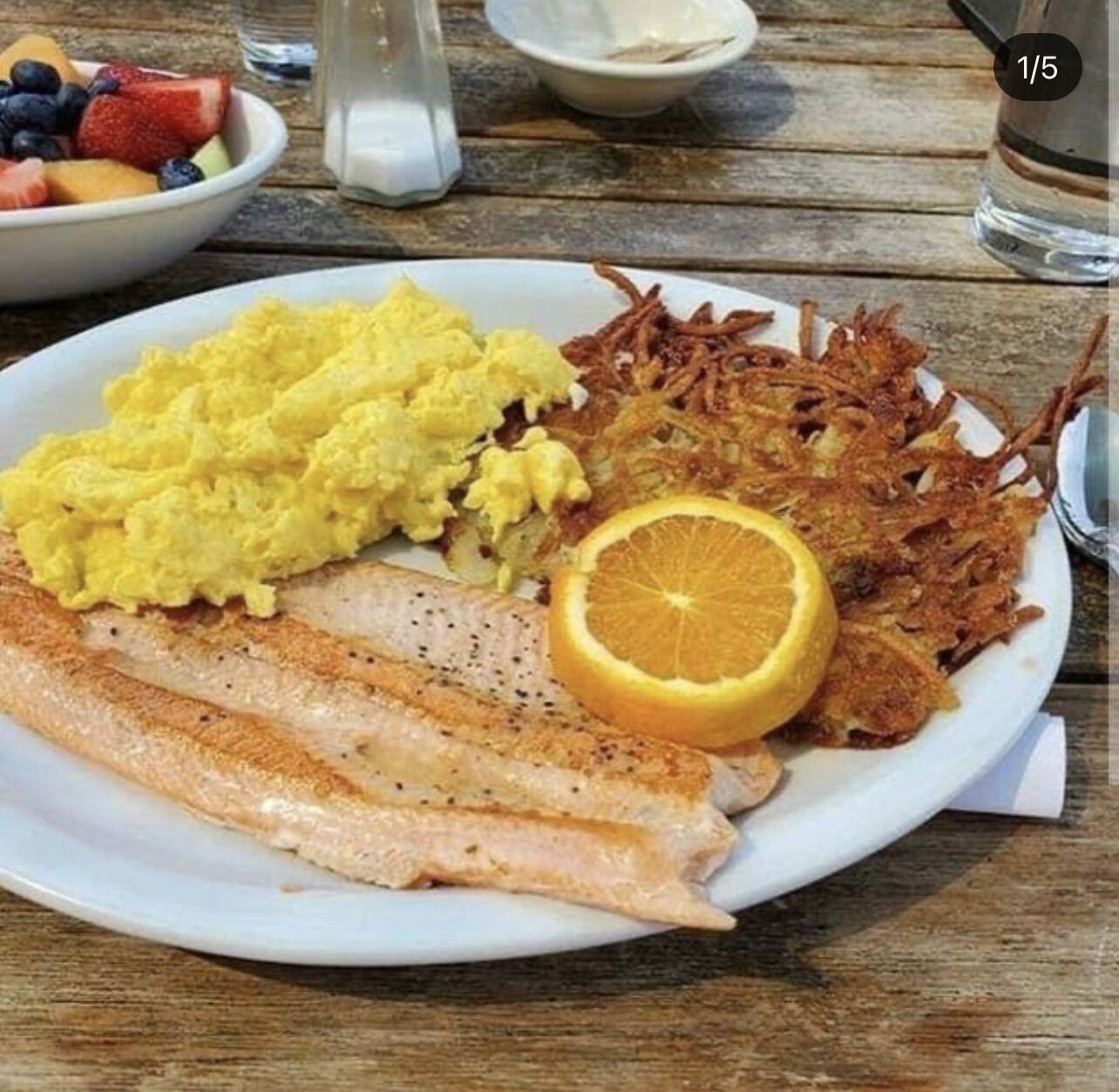 A plate with scrambled eggs, fried fish, hash browns, and a slice of orange on a wooden table.