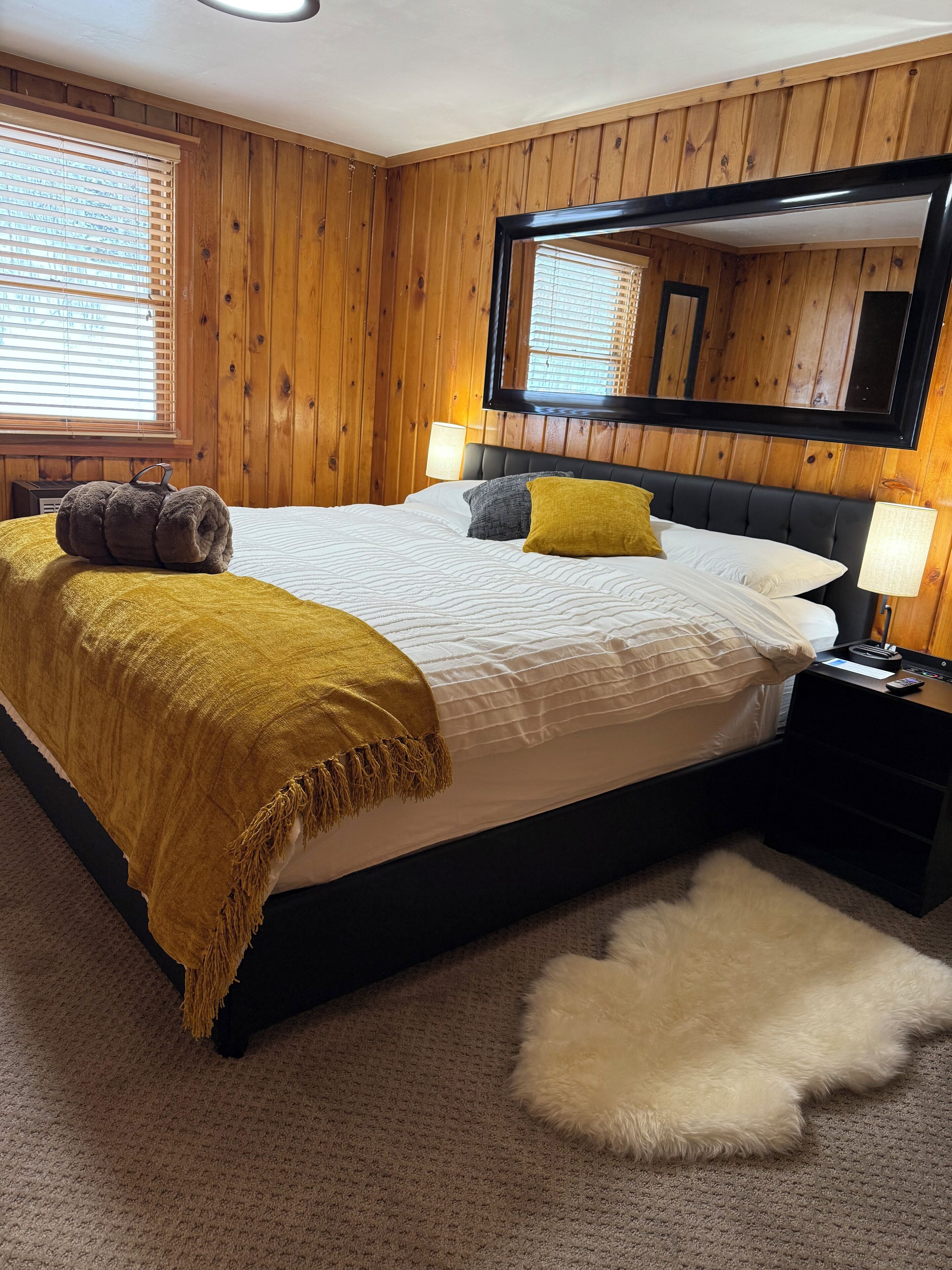 A bedroom with a wooden paneled wall, a large bed with a white comforter, two pillows, a yellow throw blanket, a fluffy white rug, and a nightstand with a lamp.