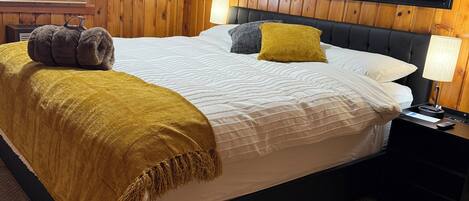 A bedroom with a wooden paneled wall, a large bed with a white comforter, two pillows, a yellow throw blanket, a fluffy white rug, and a nightstand with a lamp.