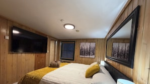 A bedroom with a large bed, a flat-screen TV, wooden paneling, a window with blinds, and a mirror. - Silver Fork Lodge and Restaurant (Brighton)