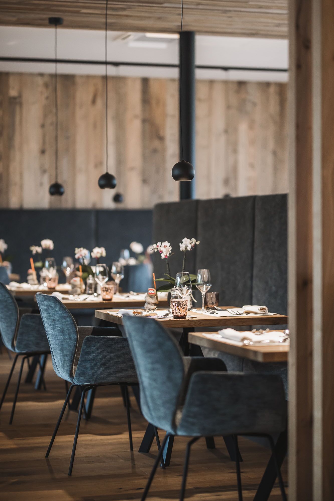Un restaurante con paredes de madera, lámparas colgantes y sillas tapizadas en azul. Las mesas están preparadas con copas de vino y pequeños jarrones.