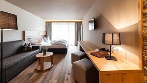 A modern hotel room with a sofa, a small round table, a desk with a lamp, a bed with a headboard, and a stone accent wall. - Autentis (Rasun Anterselva)