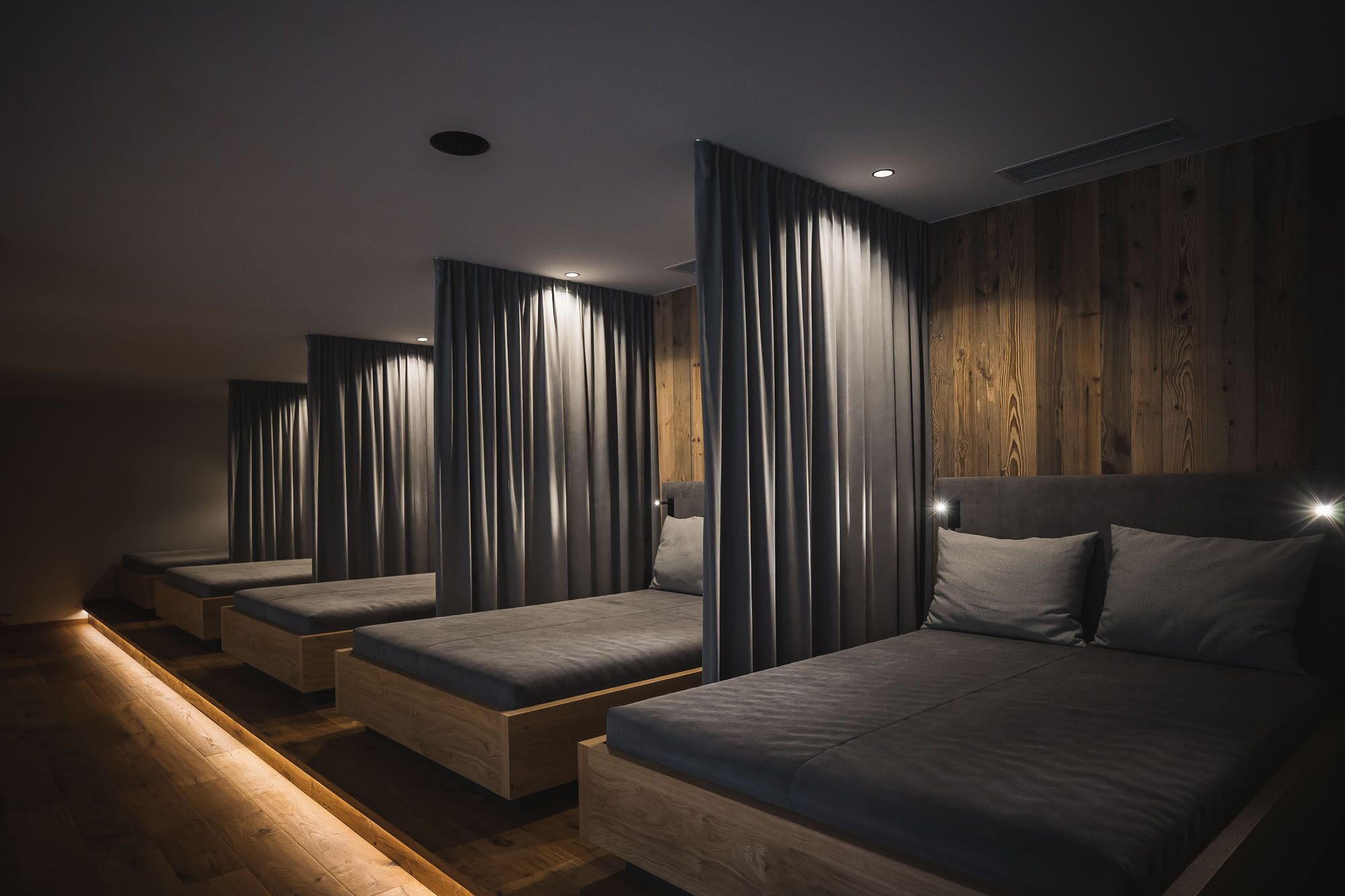 A row of four single beds with wooden headboards and grey bedding, in a dimly lit room with wooden paneling on the walls.