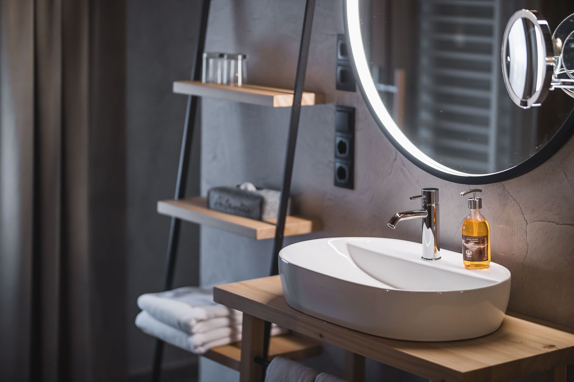 Un baño moderno con un espejo redondo, un lavabo blanco y un dispensador de jabón sobre una repisa de madera.