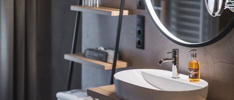 Un baño moderno con un espejo redondo, un lavabo blanco y un dispensador de jabón sobre una repisa de madera.