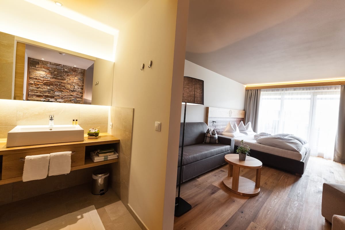 Habitación de hotel moderna con baño que incluye un espejo grande, lavamanos y una pared decorativa de piedra. El dormitorio tiene una cama, un sofá y una pequeña mesa redonda.