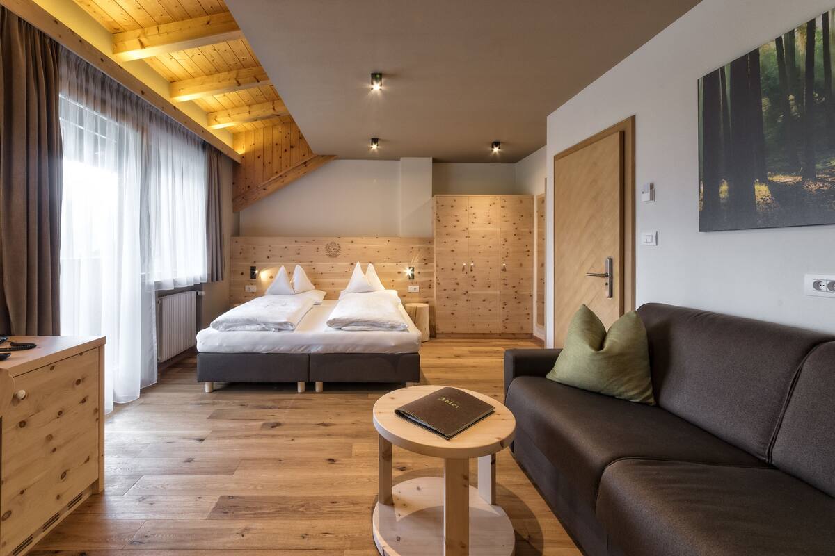 Un dormitorio moderno con techo de madera, una cama grande con ropa de cama blanca, un aparador de madera, una mesa redonda de madera y un sofá gris.