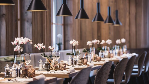 A long dining table set with chairs, black pendant lights, and floral centerpieces. - Autentis (Rasun Anterselva)