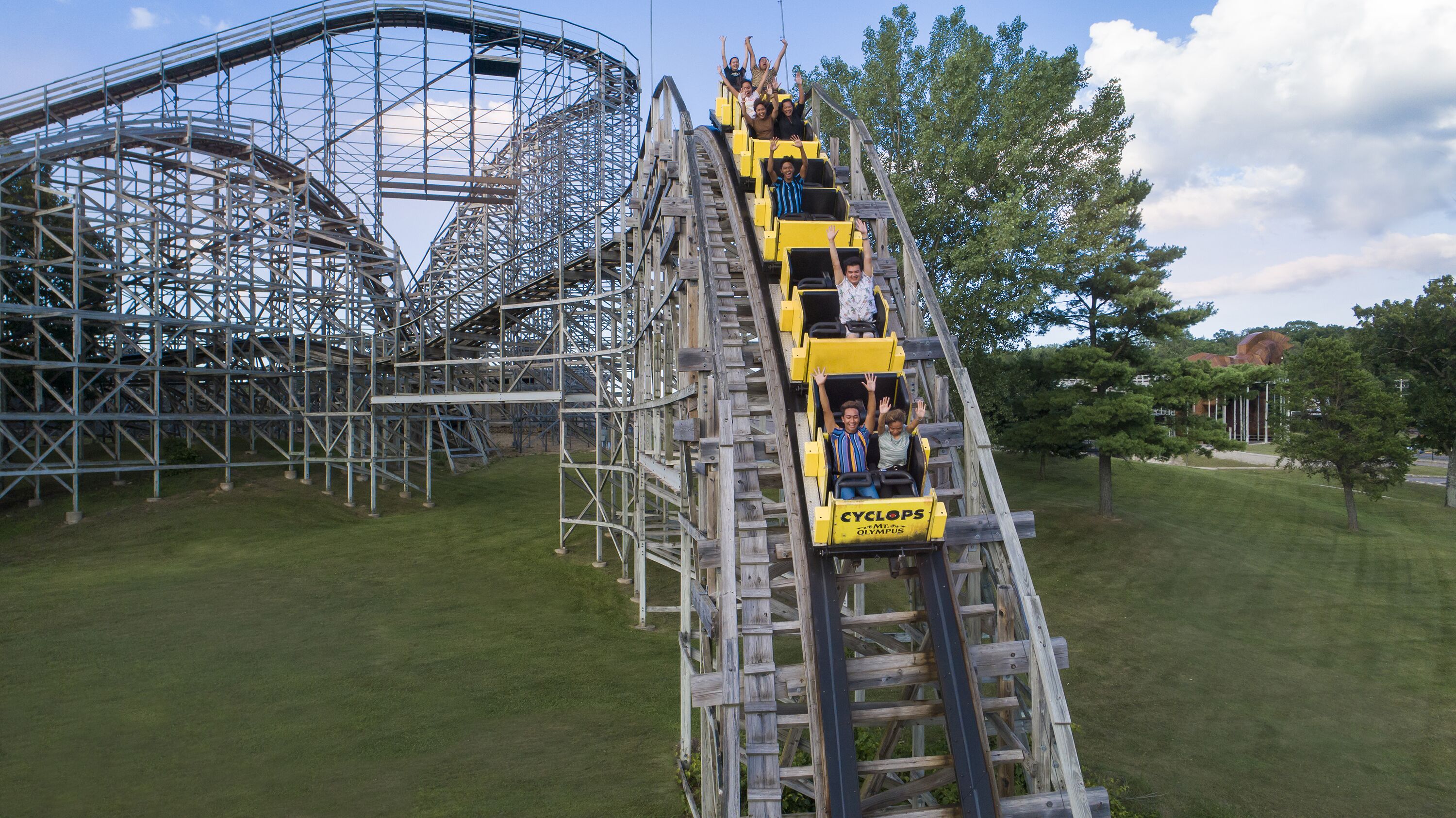 A wooden roller coaster with passengers, surrounded by metal scaffolding and trees.