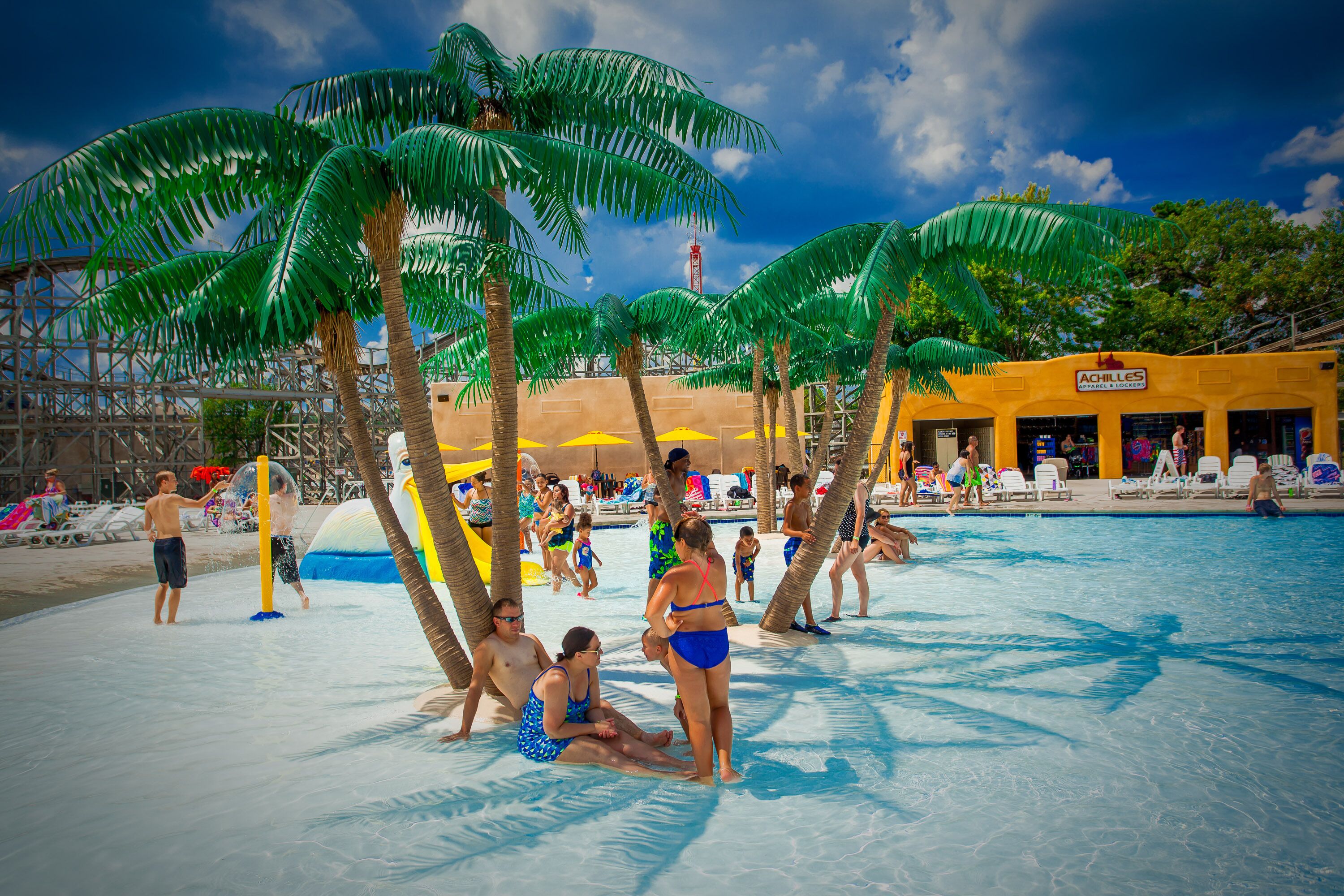 A pool area with palm trees, a yellow building, and people swimming and relaxing.