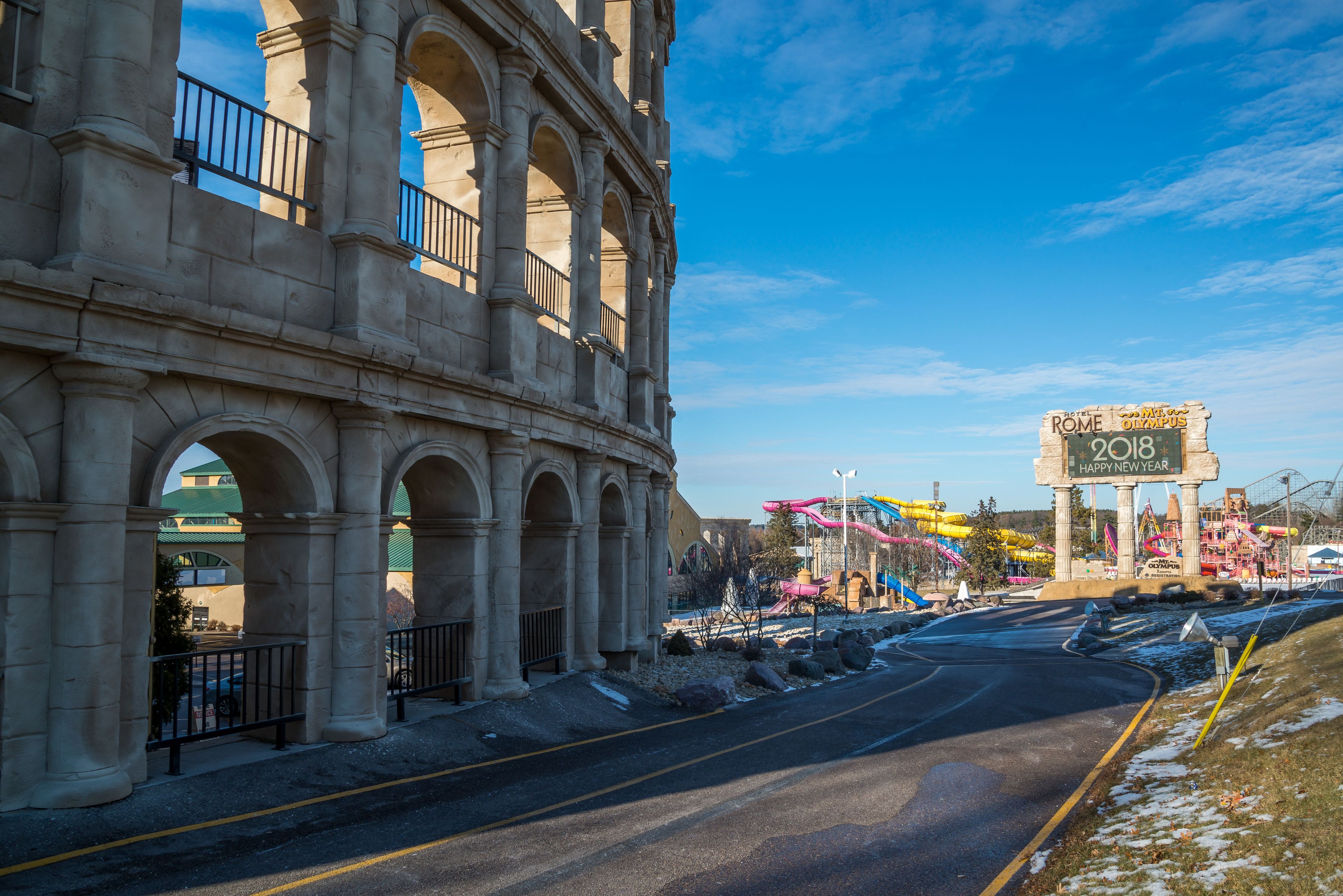 A large, historic building with multiple arches and columns, a sign reading "ROME 2018," and a road in the foreground.