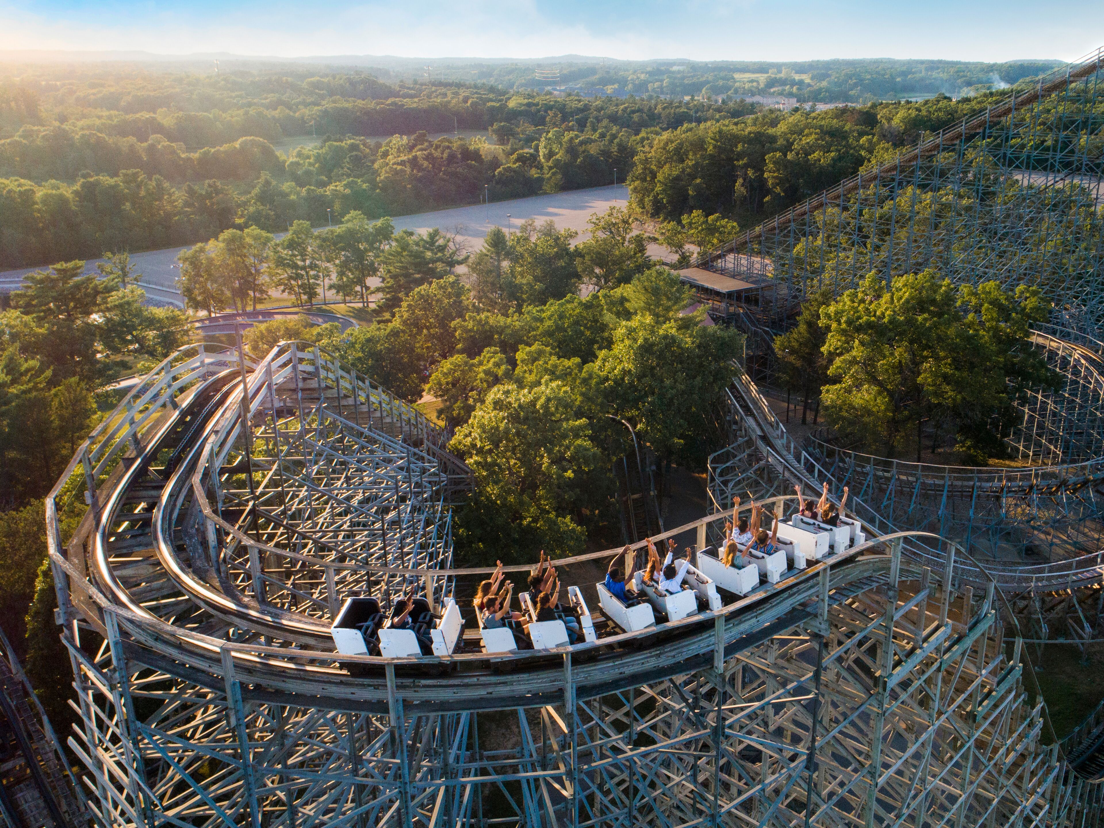 A roller coaster with multiple cars and riders, surrounded by a forested area and a river in the background.