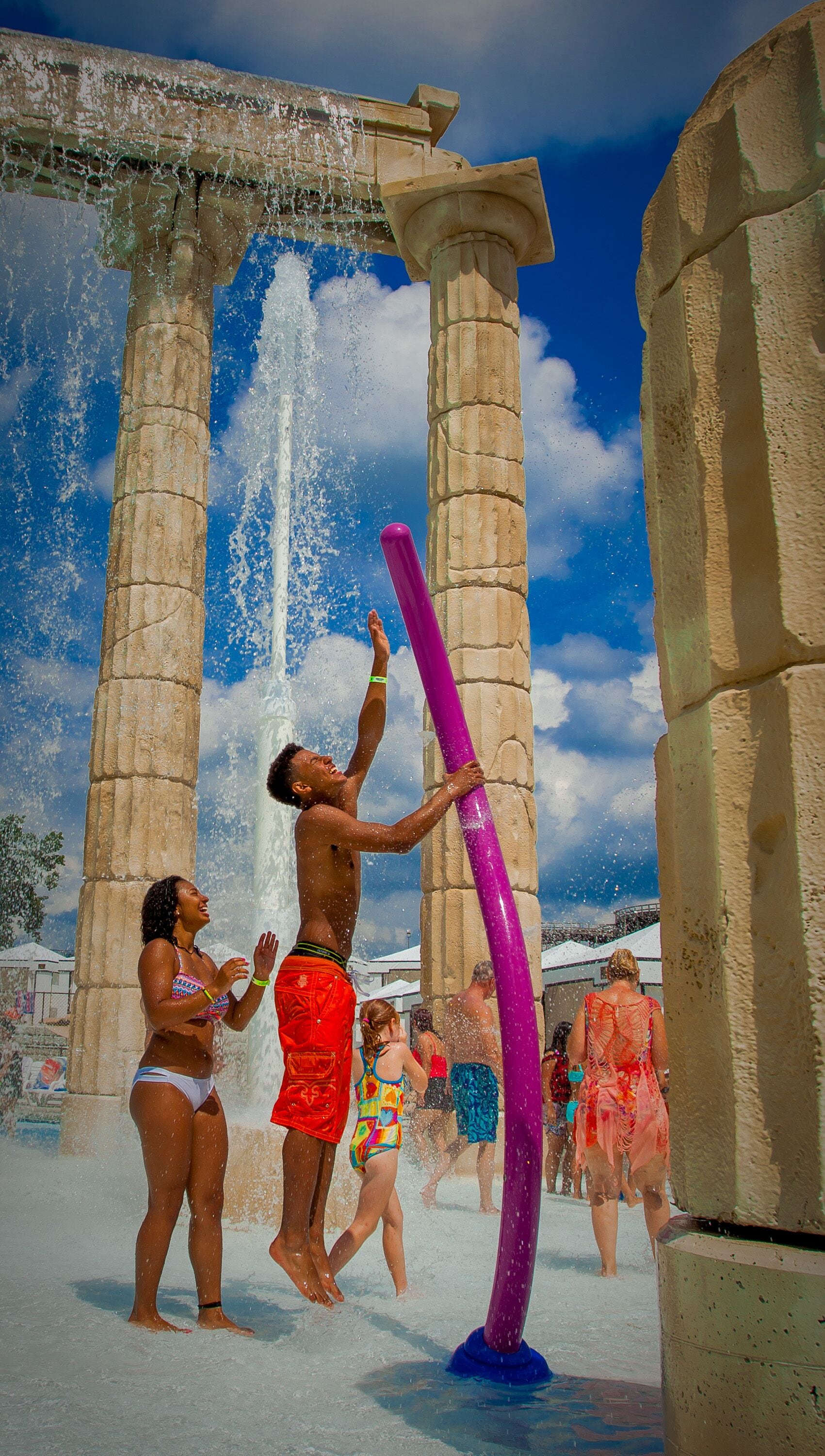 A water fountain with a column structure, surrounded by people enjoying the water.