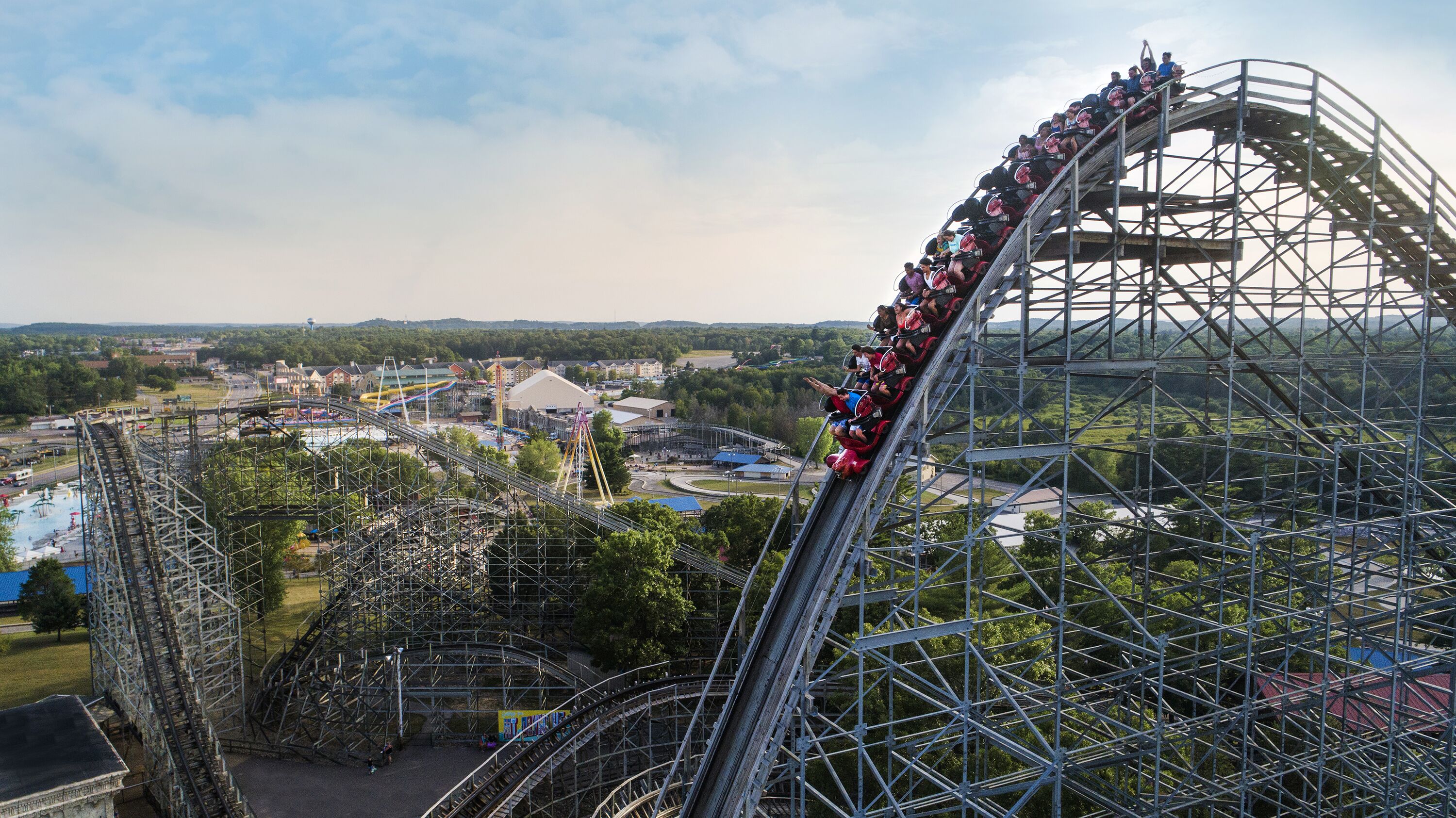 A roller coaster with a steep incline and a long track, surrounded by a park with various structures and greenery.