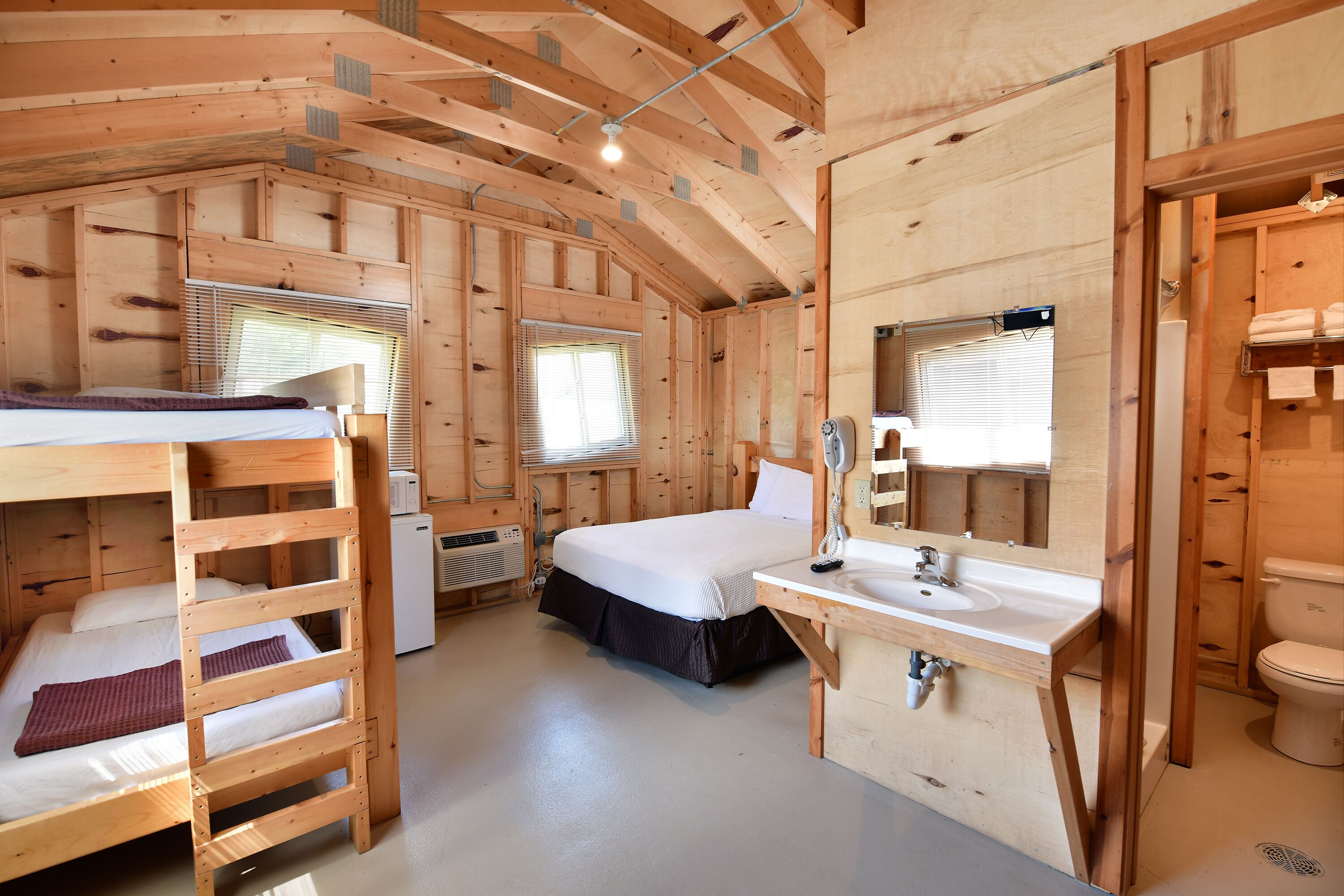 A wooden cabin with bunk beds, a single bed, a bathroom with a sink and toilet, and a window with blinds.