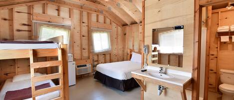 Une cabane en bois avec des lits superposés, un lit simple, une salle de bain avec un lavabo et des toilettes, et une fenêtre avec des stores.