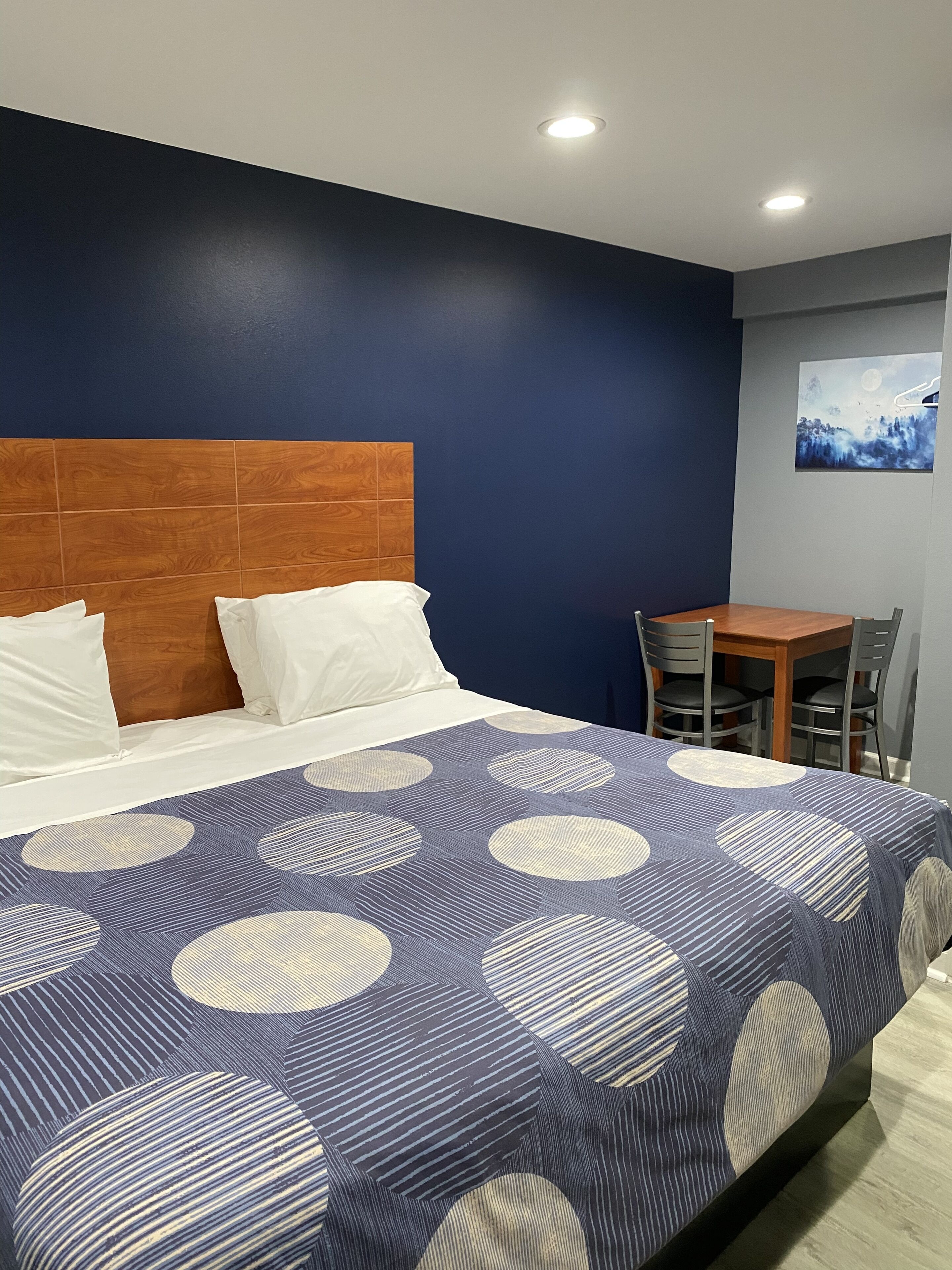 Habitación de hotel con cama, cabecero de madera, mesa de comedor con sillas y un cuadro en la pared.