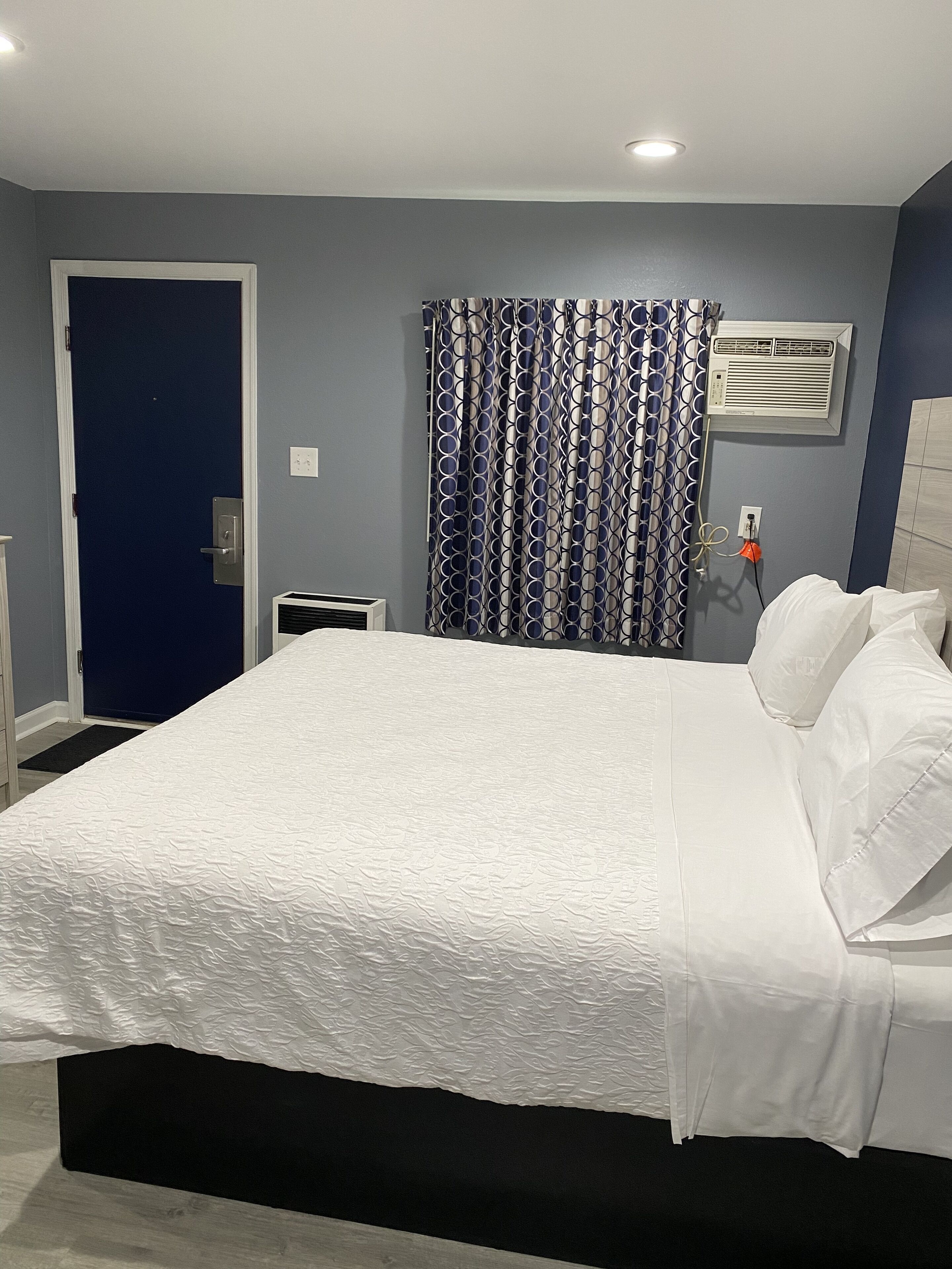 A neatly made bed in a room with a dark blue door, a patterned curtain, and an air conditioning unit on the wall.