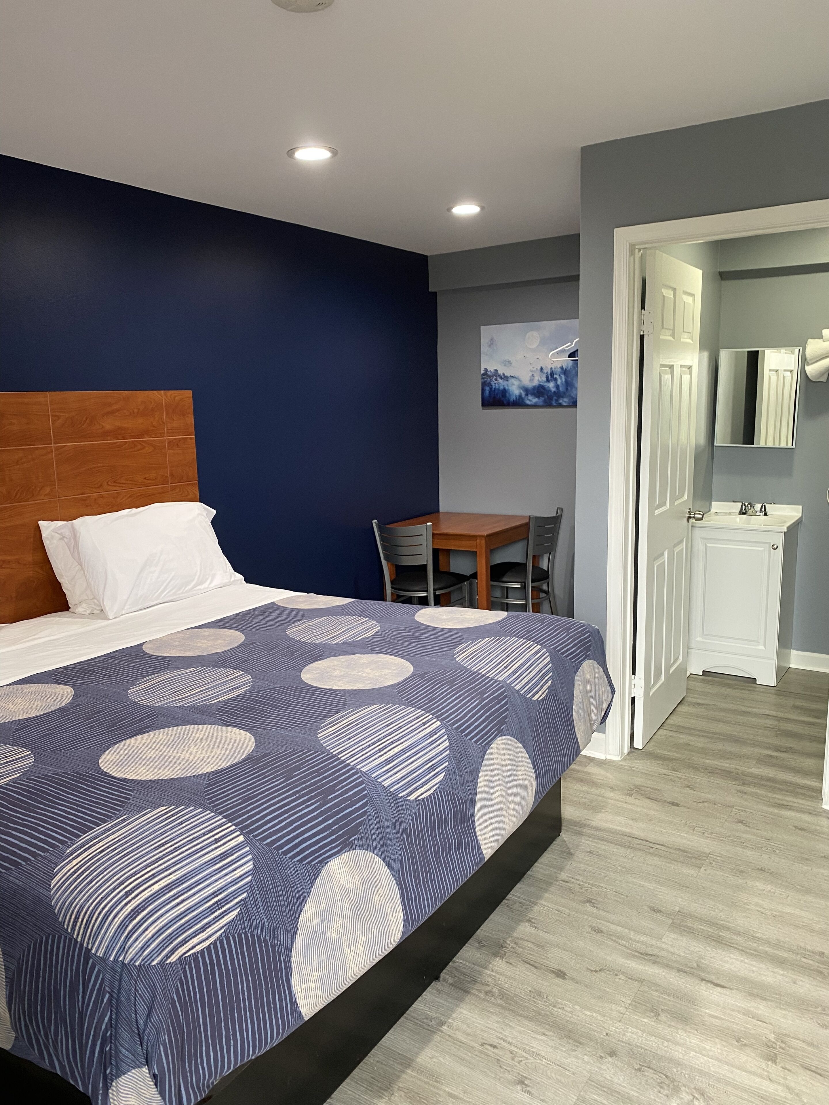 Un dormitorio con una cama grande, cabecera de madera, una mesa de comedor con sillas, un baño con espejo y lavamanos, y un cuadro en la pared.