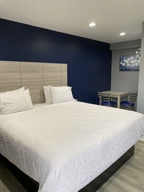 A large bed with a wooden headboard, white bedding, and a small table with chairs in a room with dark blue walls and recessed ceiling lighting.